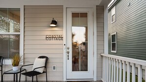 Entrada de uma casa costeira com porta branca, um pequeno alpendre com uma cadeira e mesa, e uma placa com a inscrição 'COASTAL COTTAGE'.