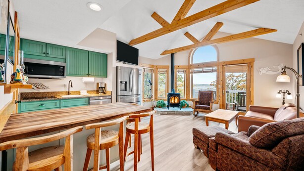 A spacious kitchen with wooden beams, a dining area with a wooden table and chairs, a cozy living room with a patterned sofa, and a fireplace. - Stony Brook 23 | Top Floor, Heated Pool, Lake Michigan View (Glen Arbor)