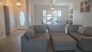 Un salon spacieux avec des canapés et des poufs gris, un coin repas avec une table et des chaises, et une vue sur l’extérieur grâce à de grandes fenêtres.