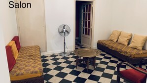 Een kamer met een zwart-wit geruite vloer, twee banken met een gedessineerde bekleding, een ventilator en een kleine ronde tafel.