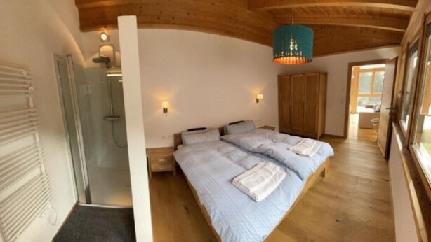 Une chambre avec un plafond en bois, un lit recouvert de draps blancs, une suspension bleue et une armoire en bois.