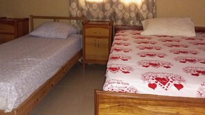 A bedroom with two single beds, a wooden nightstand, and a window with patterned curtains. - Darsalam (Kedougou)