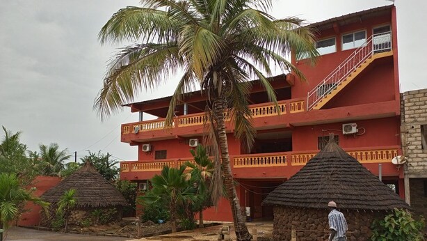 A multi-story building with a red facade, a balcony, and a thatched-roof structure in the foreground. - Darsalam (Kedougou)