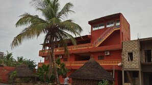 A multi-story building with a red facade, a balcony, and a thatched-roof structure nearby. - Darsalam (Kedougou)