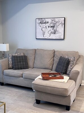 A living room with a grey sofa, patterned cushions, a wooden tray, and a framed inspirational quote on the wall.