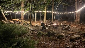 Een terras met houten banken en stoelen, omringd door bomen en verlicht met lichtslingers.
