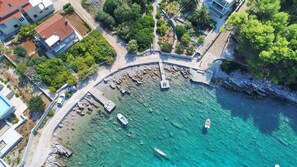 Vista aérea de una zona costera con aguas turquesas cristalinas, costas rocosas y varios barcos anclados. Se observan edificios con tejados de baldosas rojas y un paseo marítimo pavimentado.