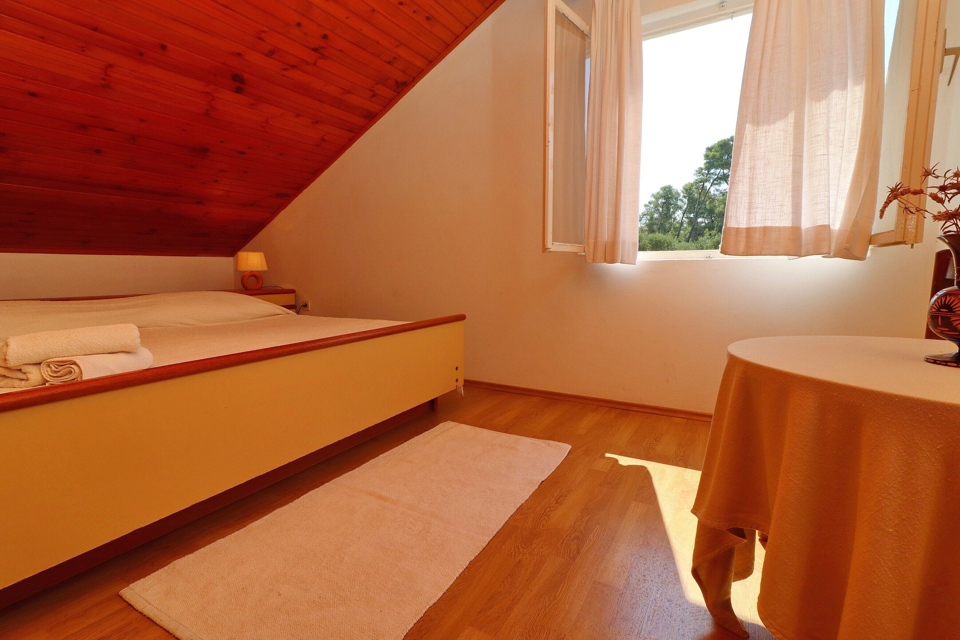 Un dormitorio con techo de madera, una cama, una mesita de noche con lámpara, una ventana con cortinas y una mesa redonda con mantel.