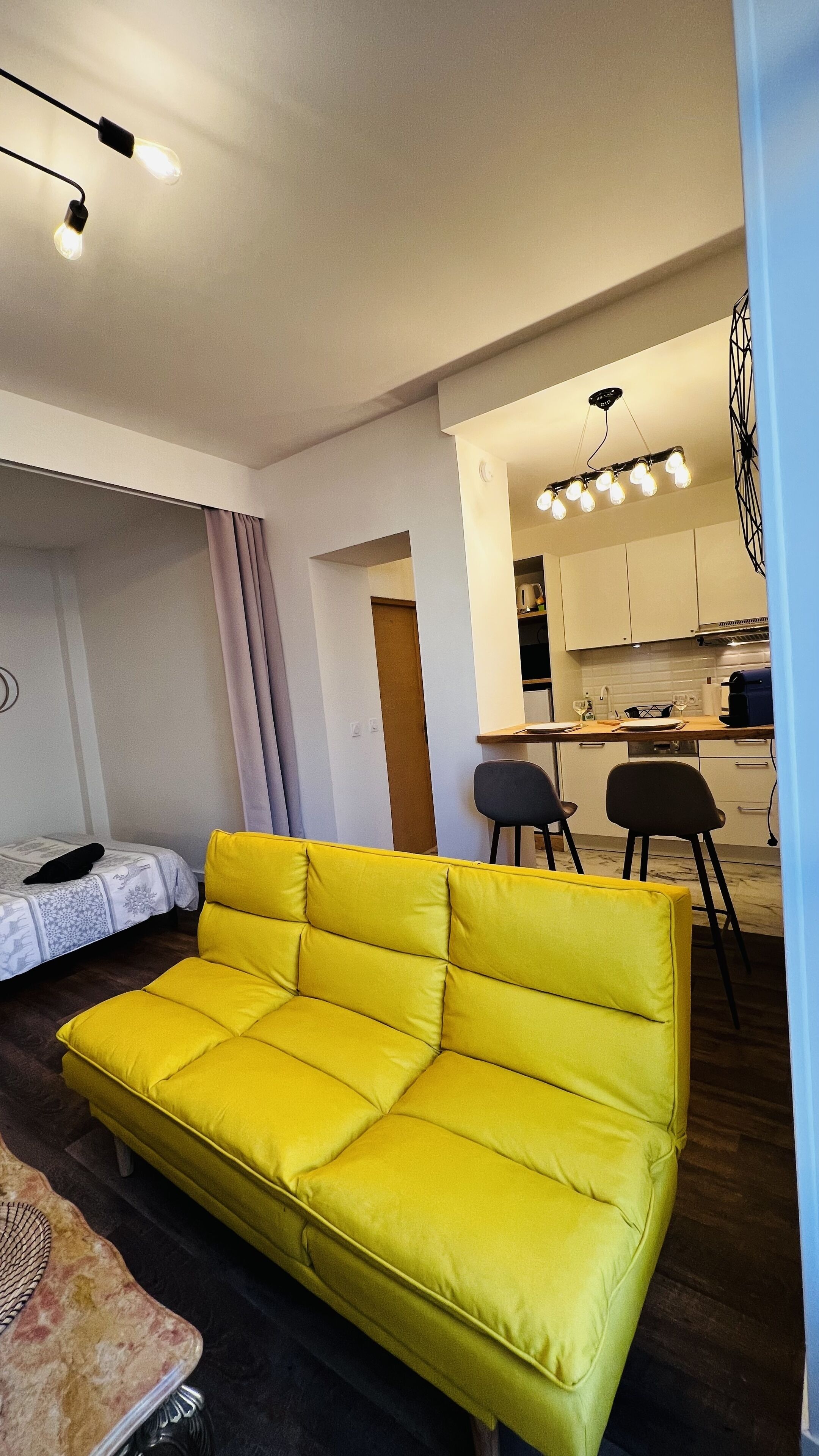 A modern living room with a bright yellow sofa, a small dining area with two chairs, and a kitchen in the background.