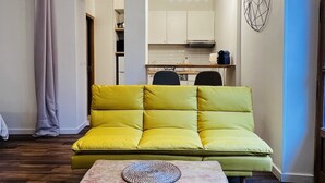Un salon moderne avec un canapé jaune, une table basse sur laquelle repose un dessous de verre en fibres tressées, et une cuisine en arrière-plan.