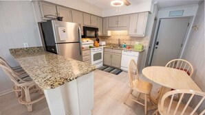 Una cocina con encimeras de granito, muebles de cocina blancos, un refrigerador de acero inoxidable, un microondas y una estufa. Hay una zona de comedor con una mesa y sillas de madera.