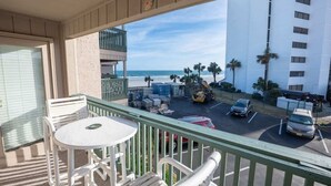 Un balcón con vista a la playa, un estacionamiento con vehículos y un edificio al fondo.