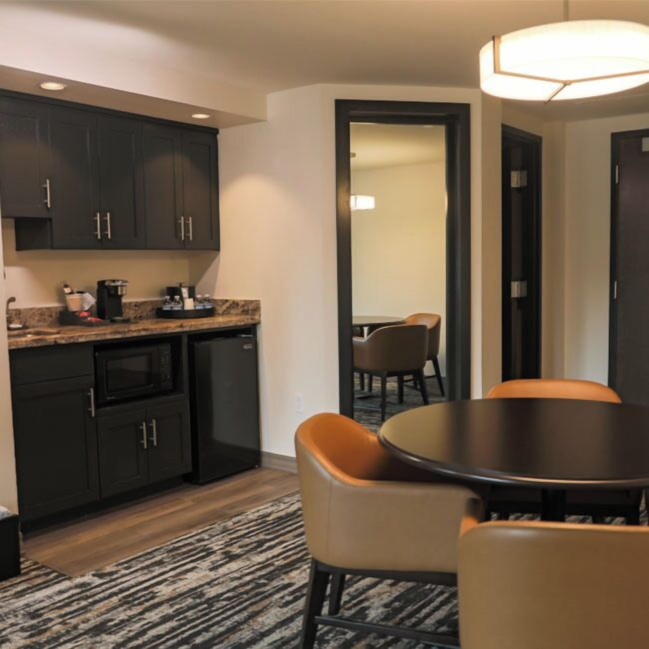 A compact kitchen with dark cabinetry, a built-in microwave, and a coffee maker. There is a round table with two chairs and a patterned rug on the floor.