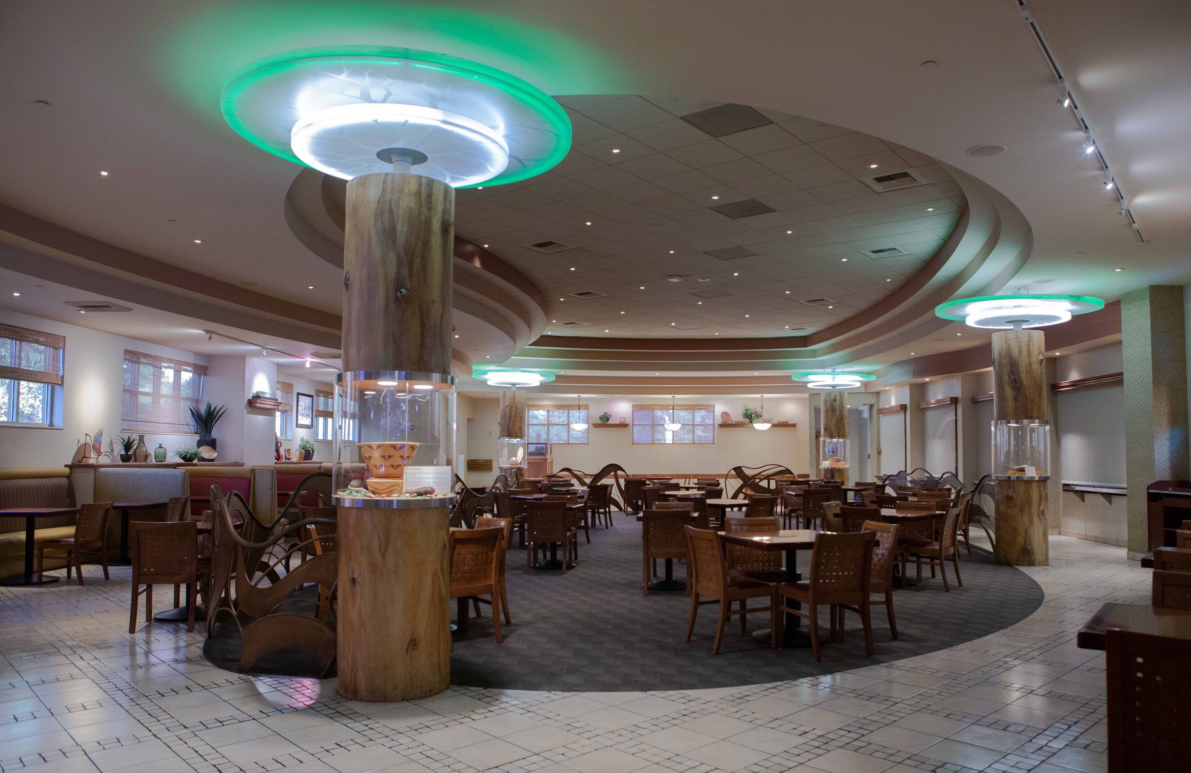Un restaurant spacieux avec des tables et des chaises en bois, de grandes colonnes en bois et un éclairage d’ambiance vert.