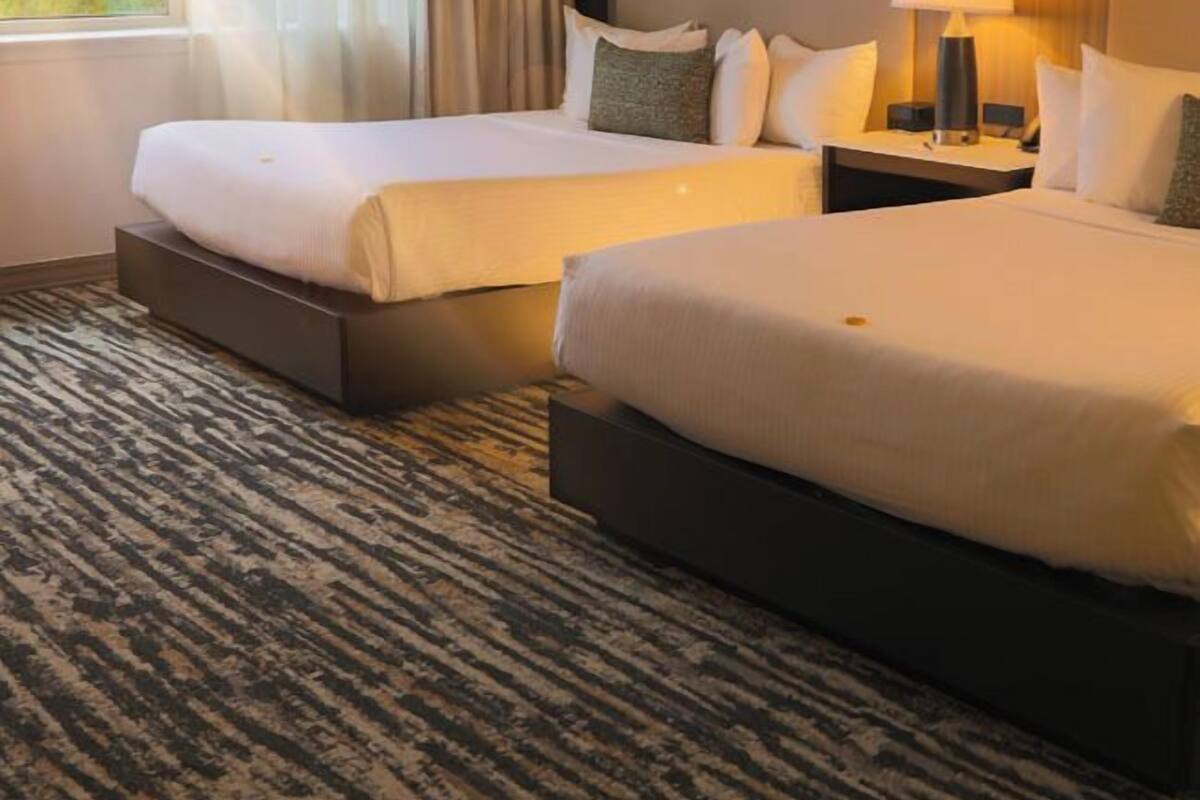 A hotel room with two beds, a large window with curtains, a nightstand with a lamp, and a patterned carpet.