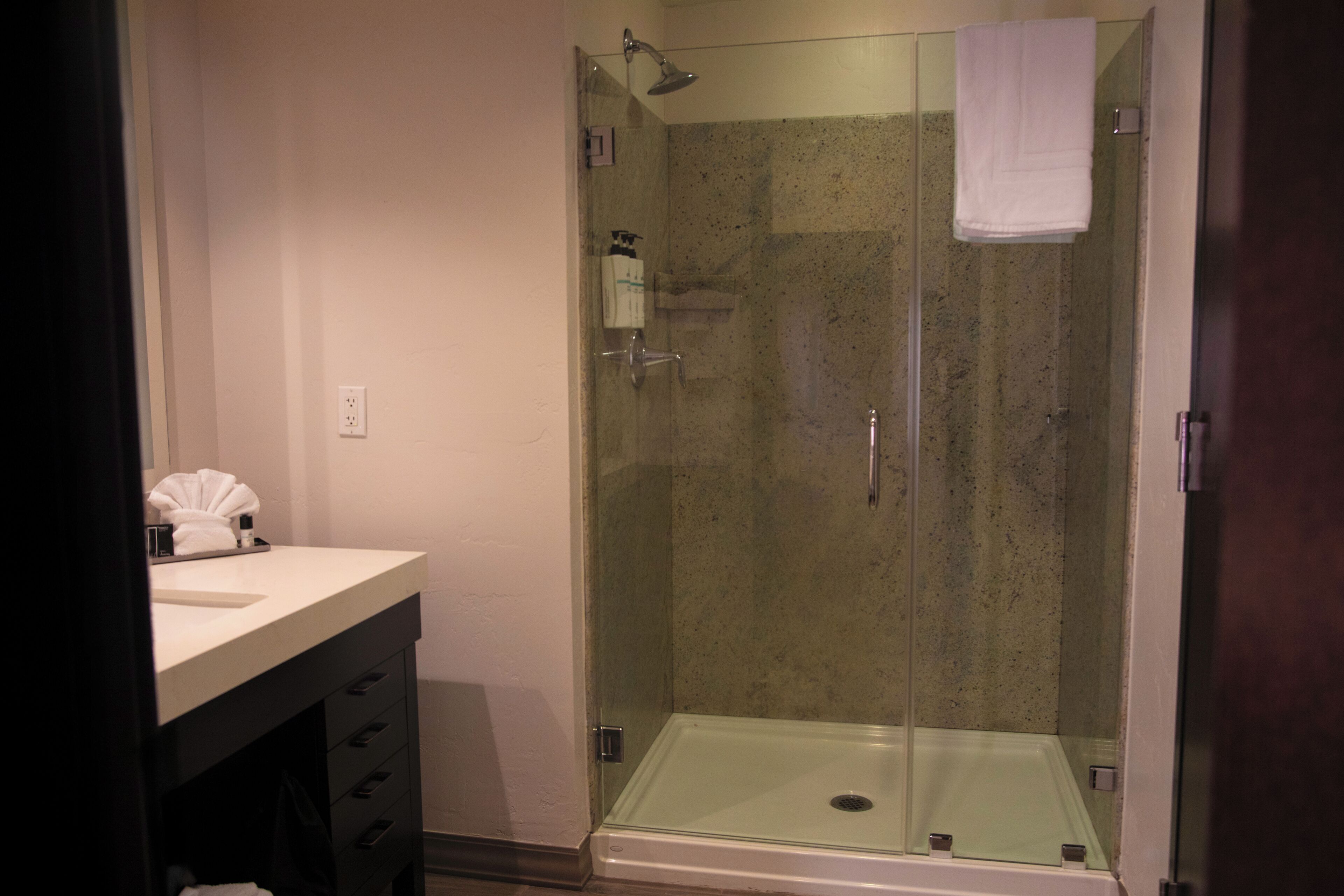 A glass-enclosed shower with a white towel hanging on the wall, a sink with toiletries, and a dark-colored cabinet.