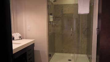 A glass-enclosed shower with a white towel hanging on the wall, a sink with toiletries, and a dark-colored cabinet.