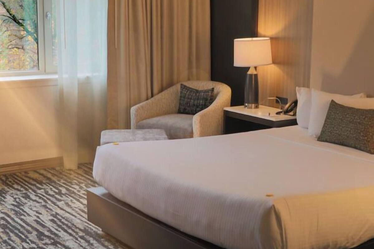 A hotel room with a large bed, a chair, a nightstand with a lamp, and a window with curtains.