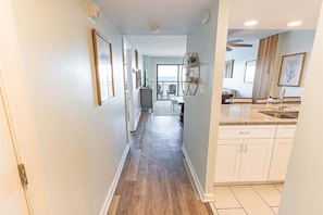 A narrow hallway with wooden flooring, white walls, and a kitchen area with white cabinets and a sink.