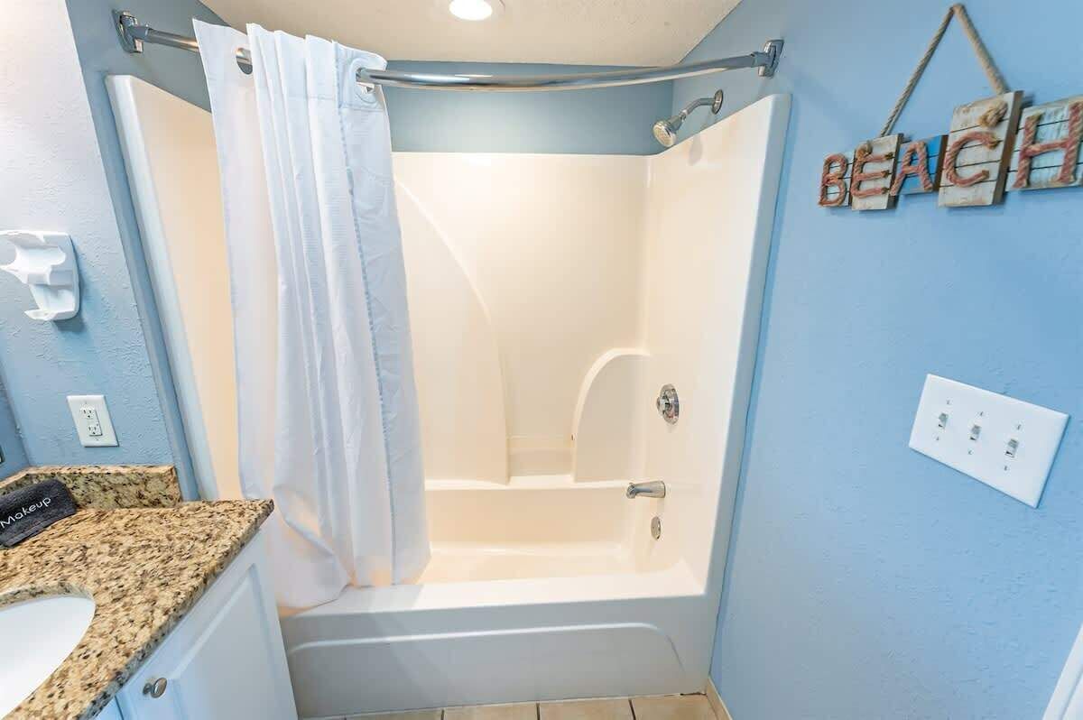 A bathroom with a shower, a towel rack, a sink with a granite countertop, and a wall-mounted light switch.