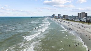 Een strand met golven, een pier en gebouwen op de achtergrond.