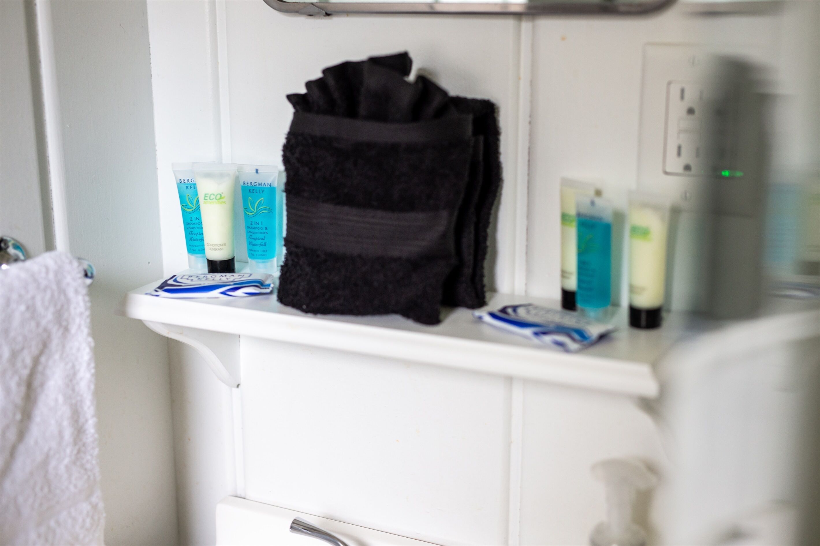 Une étagère de salle de bain contenant des articles de toilette, notamment une serviette noire, un shampoing Bergman Kelly et d’autres produits d’hygiène personnelle.