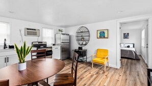 Una cocina moderna con armarios blancos, una mesa de comedor y una silla amarilla. En la habitación contigua hay una cama con una colcha gris.