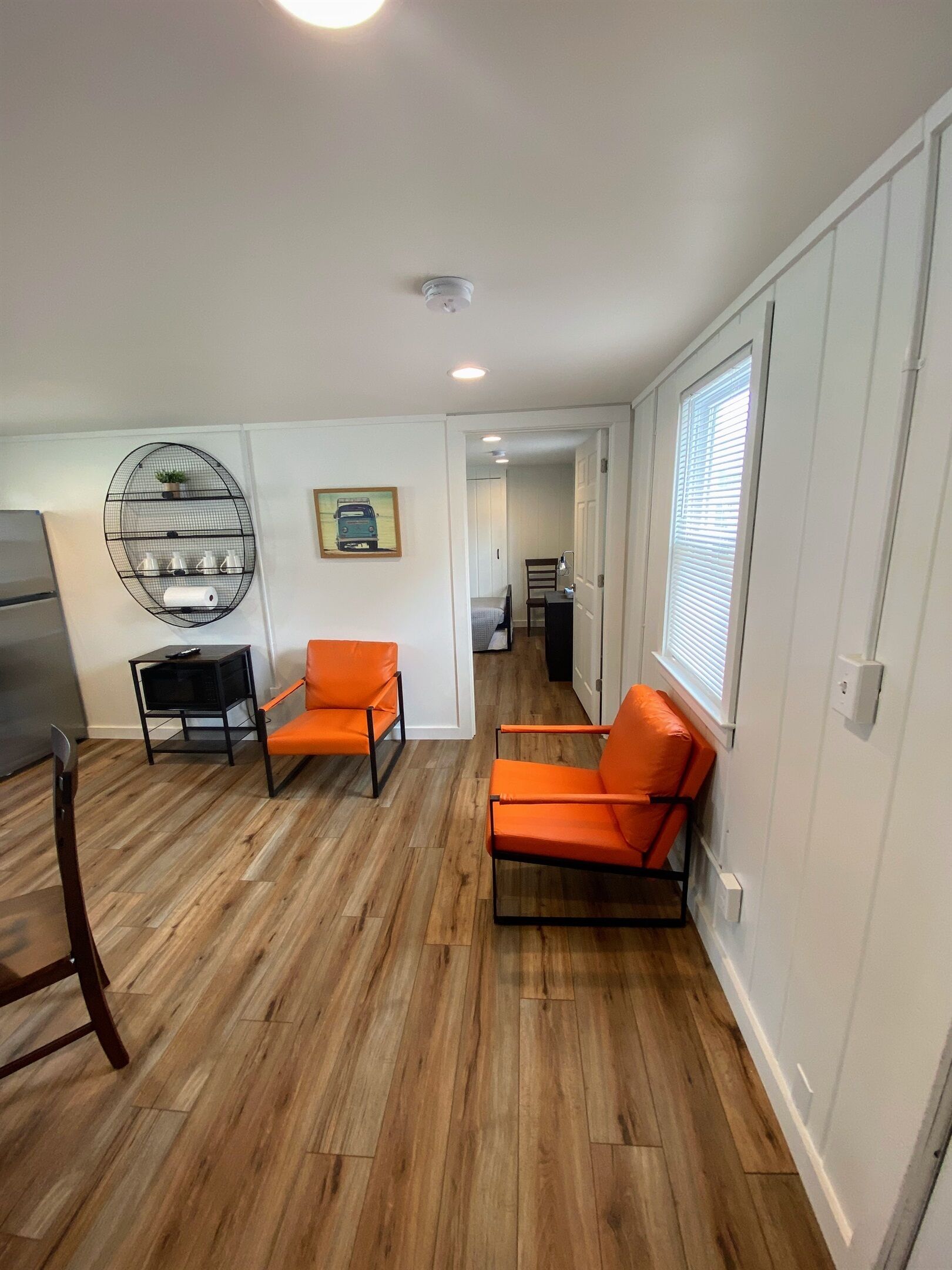 A spacious living room with hardwood flooring, two orange chairs, a round wire shelf with decorative items, and a view into the kitchen area.
