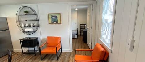 A spacious living room with hardwood flooring, two orange chairs, a round wire shelf with decorative items, and a view into the kitchen area.