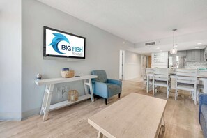 A modern living room with a television displaying 'BIG FISH Rentals,' a blue armchair, a white dining table, and a wooden coffee table. - Amazing North Tower at Sand Dunes Resort (Myrtle Beach)