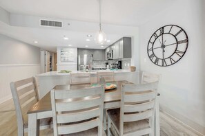 A modern kitchen with a dining area, featuring a large clock on the wall, wooden chairs, and a central island. - Amazing North Tower at Sand Dunes Resort (Myrtle Beach)