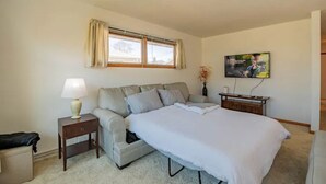 A bedroom with a bed, a nightstand, a lamp, a window with curtains, a TV, and a small table with a vase.