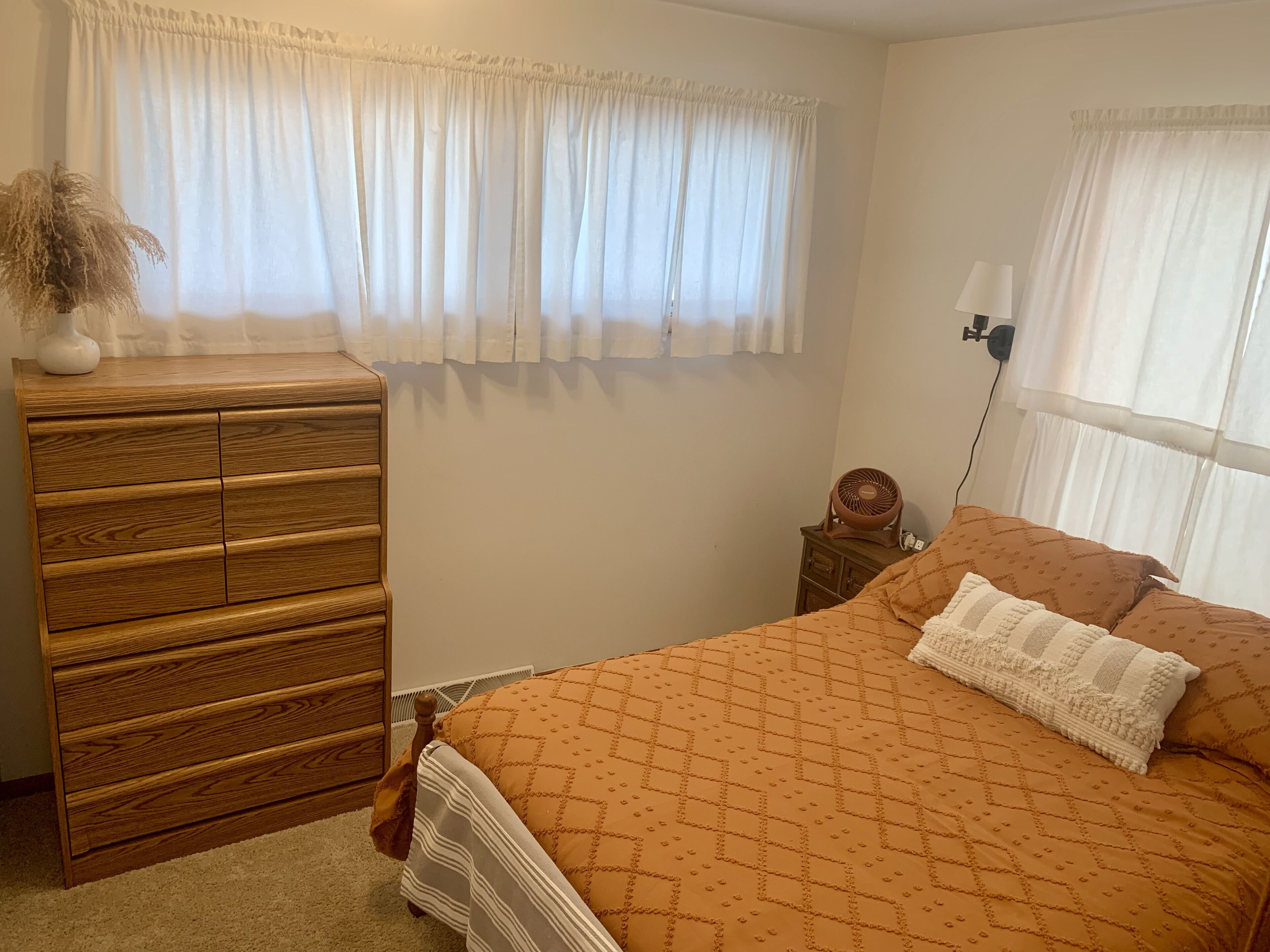 A bedroom with a wooden dresser, a bed with an orange patterned bedspread, and a window with white curtains.