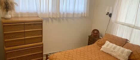 A bedroom with a wooden dresser, a bed with an orange patterned bedspread, and a window with white curtains.