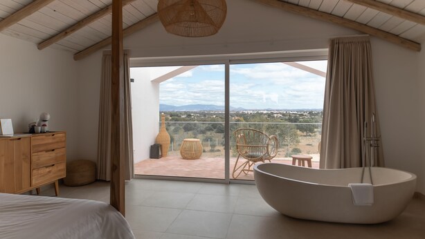 Quarto com cama grande, banheira, cadeiras de vime e vista para o campo.