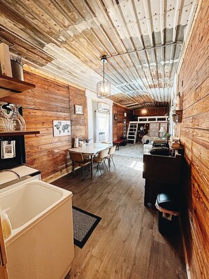 A rustic interior with wooden walls, a dining area with chairs, a kitchen with a sink, and a ladder leading to a garage.