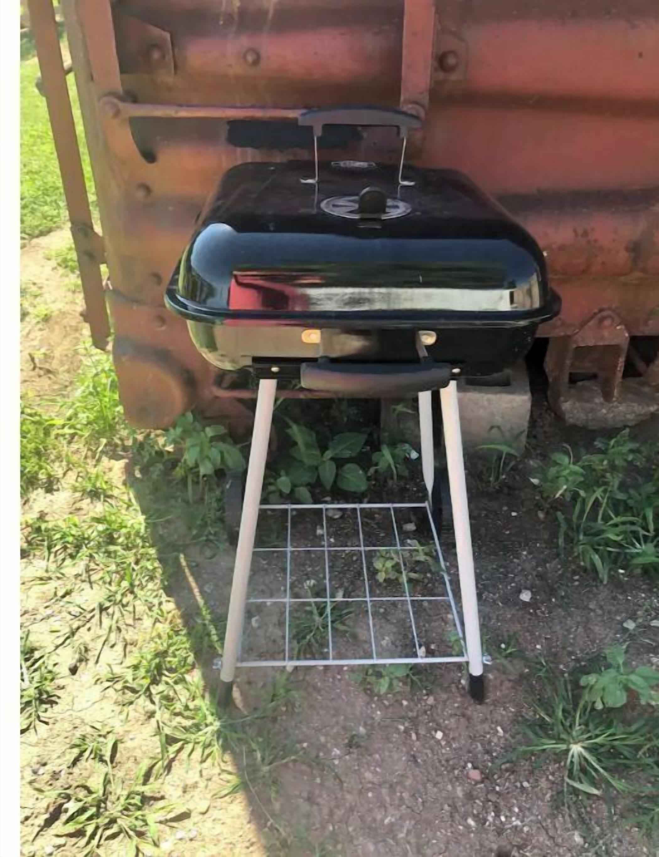 A portable grill with a metal grate and a handle, placed on a dirt ground with sparse vegetation.