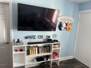A living room with a TV mounted on the wall, a bookshelf with books, a sign reading "Beach House", and a door. - Beachfront condo with spectacular views - steps from the beach (Wildwood Crest)