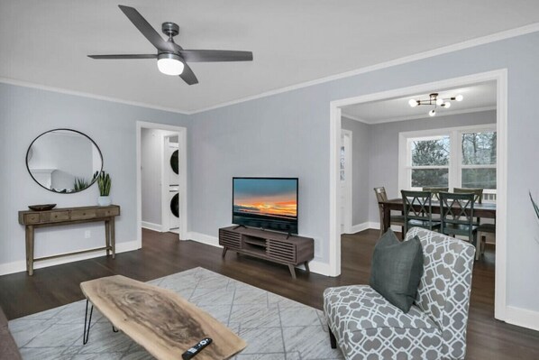 A modern living room with a flat-screen TV, a ceiling fan, a dining area with a table and chairs, and a large window. - Pristine 3BR | Centrally located & Pet Friendly (Columbia)