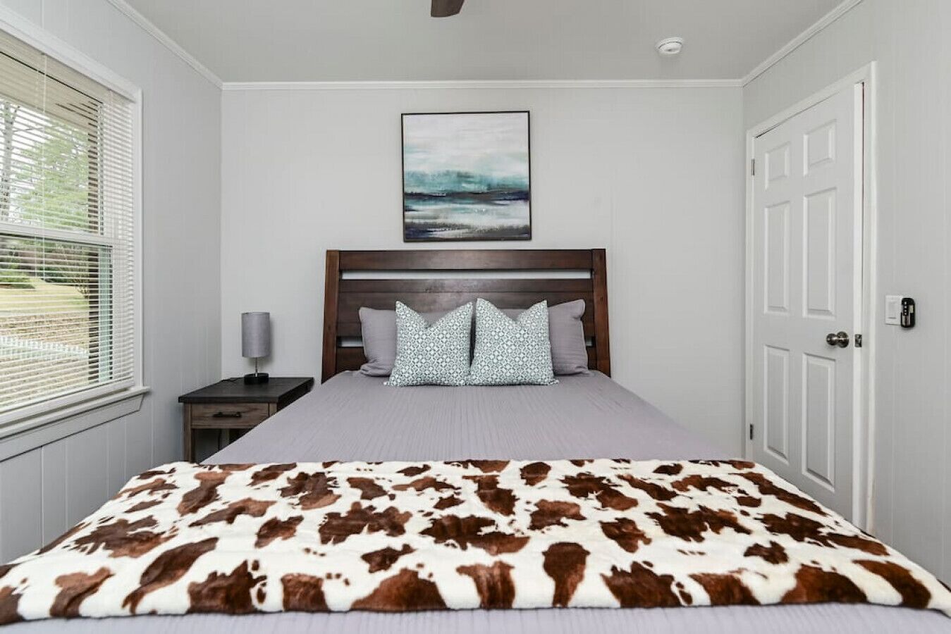 A bedroom with a bed, a nightstand, a lamp, a door, and a window with blinds.