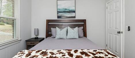 A bedroom with a bed, a nightstand, a lamp, a door, and a window with blinds.