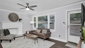 Una sala de estar con sofá, sillón y mesa de centro, chimenea y ventana grande con vista a un camino de entrada y árboles.
