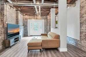 A modern living room with exposed brick walls, a large flat-screen TV, a beige sofa, and a wooden floor. - Pristine 3BR | Centrally located & Pet Friendly (Columbia)