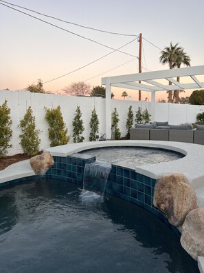 A swimming pool with a waterfall feature, surrounded by a white wall and small trees.