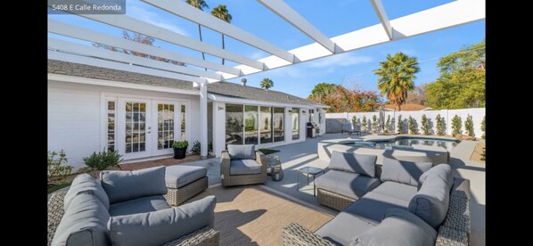 A covered outdoor patio with a pool, seating area, and a white building with large windows.