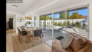 Ein modernes Wohnzimmer mit einem braunen Ledersofa, einem Holzesstisch mit Stühlen und einer großen Glastür, die zum Poolbereich führt.