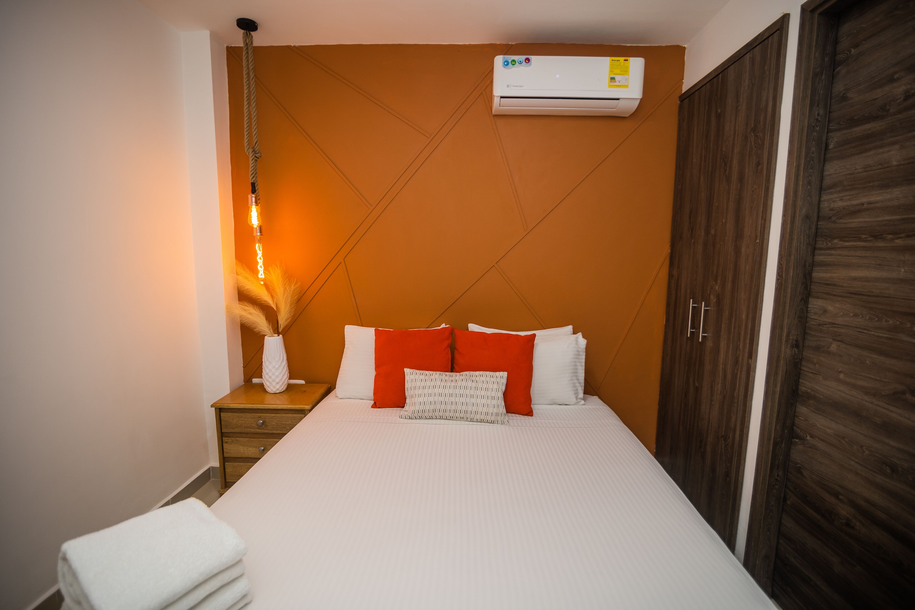 Una cama bien hecha con ropa de cama blanca y roja, una lámpara de noche y un equipo de aire acondicionado en la pared.