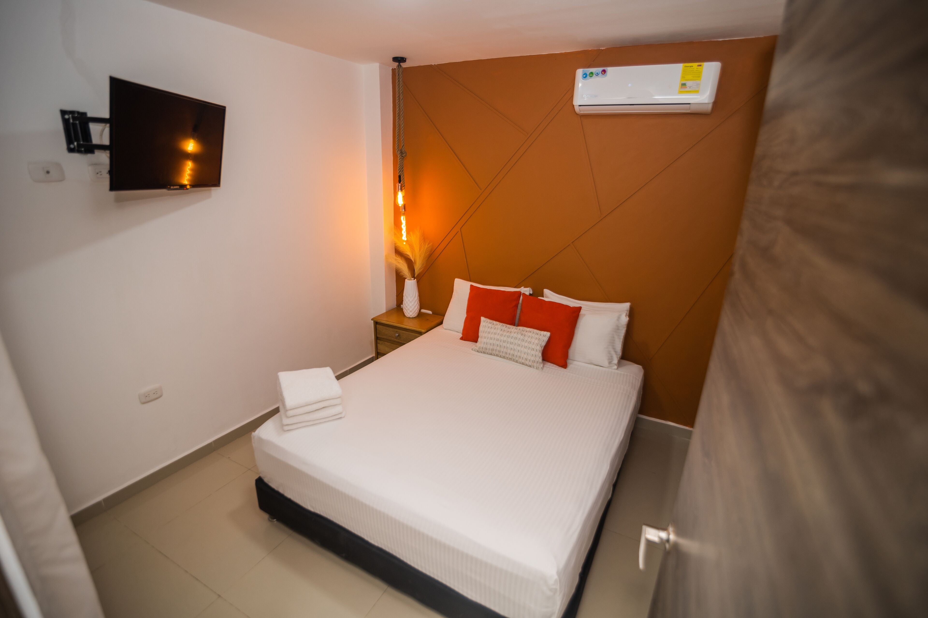 Un lit bien fait, avec une literie blanche et des oreillers orange, un téléviseur à écran plat fixé au mur et un appareil de climatisation placé au-dessus du lit.
