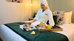 Eine Person in einem weißen Bademantel sitzt auf einem Bett und hält ein Frühstücksbrett mit Kaffee und Orangensaft.