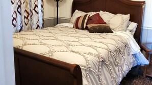 Una cama bien hecha con cabecera de madera, un plaid estampado y almohadas decorativas. Se ve también una lámpara de pie y una obra de arte en la pared.